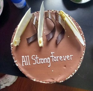 A photo of a chocolate cake seen from above. It's decorated with white chocolate discs, dark chocolate flourishes and the words "All Strong Forever" in piping.
