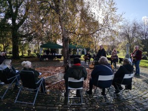 A photo of a group of musicians performing in a cemetery. The sky is blue and they are backed by trees.