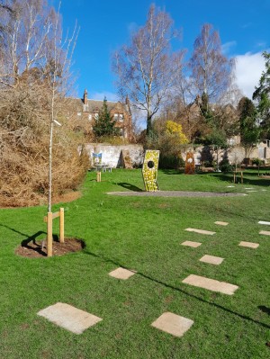 A photo of the Good Grief Garden at Dundee Botanic Gardens. Green grass, small trees, decorative stone structures and a bright blue sky.