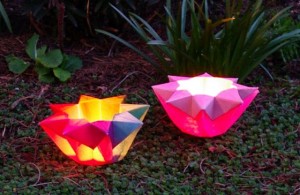 A photo of two origami paper lanterns, one red and yellow and one pink. They are lit from inside and the background is dark so they glow.