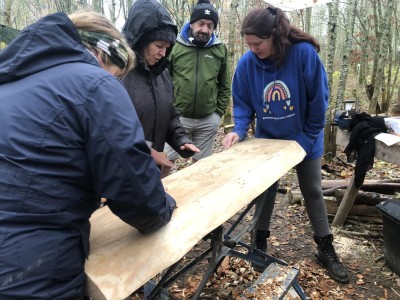 A photo of four people in a forest lifting a newly-carved piece of wood off a workbench. One is wearing a blue Borders Recovery hoodie with a rainbow logo.