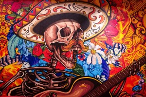 A brightly-coloured painting of a calavera with a sombrero and mustache singing and strumming a guitar against a floral background.
