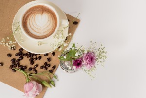 A close-up photo looking down on a cappuccino in a white porcelain cup with dried flowers around it and a brown paper napkin.