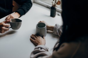 A close-up photo showing two silhouetted figures having coffee together. The photo only shows their hands, the coffees, and the top of the table.