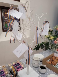Photo of a refreshments table with a grey spotted tablecloth and vase of flowers, muffins and cakes, and a memory tree with cards with names on them tied to it with ribbons.