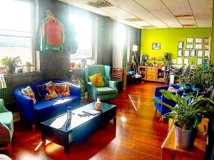 A photo of a bright, sunny seating area with a coffee table and plants. The walls are green, the floor is warm wood, and the chairs are bright blue and look comfortable. There are no people in the photo.
