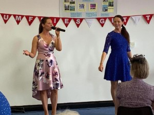 Photo of Jayd and Jenna in 50s-style dresses performing with microphones before a crowd. Behind them is a bunting that reads "Dumfries Get Together".